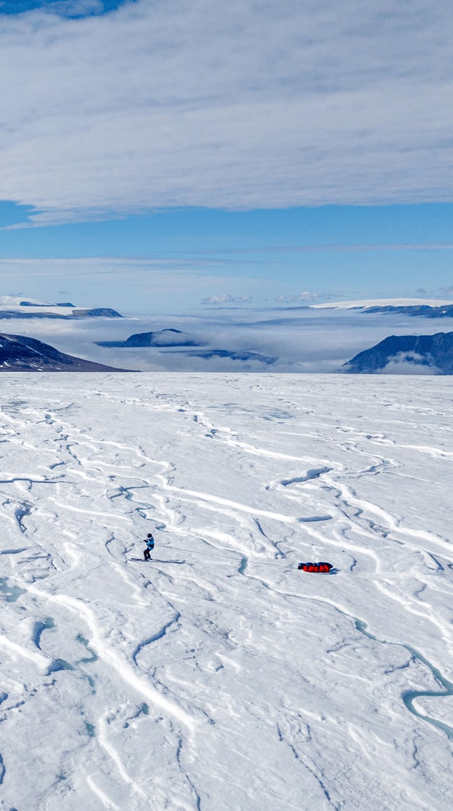 Sur une vaste étendue glacée parcourue de crevasses bleutées, Matthieu snowkite au premier plan, tiré par une voile bleue. Sa pulka rouge repose sur la glace derrière lui. En arrière-plan, on distingue les reliefs montagneux du Groenland, partiellement couverts de neige, et des nuages qui s'étendent au-dessus de la vallée. Le ciel est bleu, légèrement voilé, et la scène dégage une impression de grandeur et d'isolement au cœur d'un environnement arctique spectaculaire.