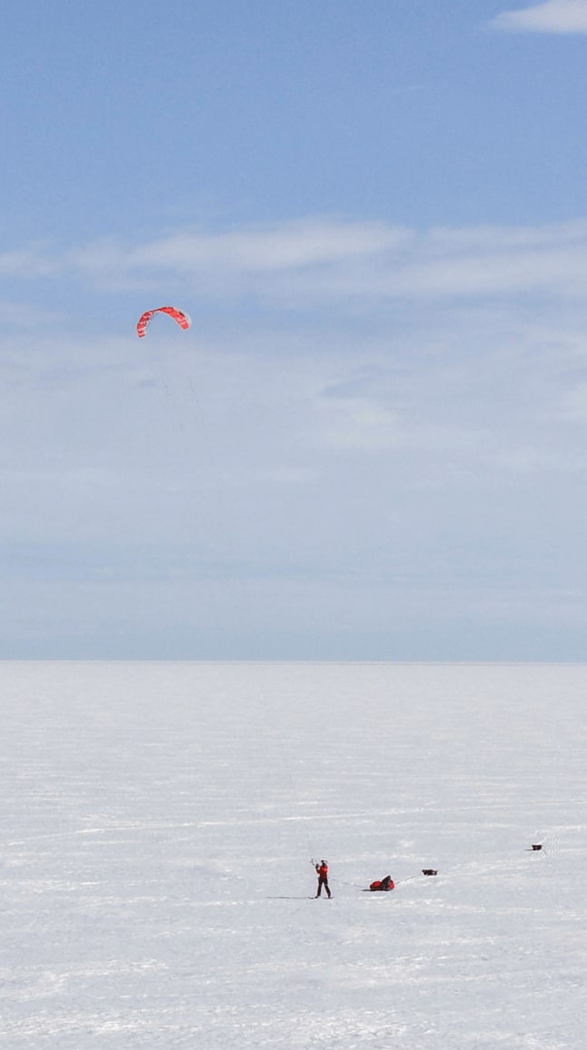 Sur une immense étendue de neige blanche, Matthieu et Heïdi avancent en snowkite sous un ciel bleu légèrement nuageux. Leurs voiles rouges se détachent dans le ciel, formant deux arcs colorés au-dessus du désert glacé. Chacun tracte une pulka rouge chargée de matériel, glissant sur la surface immaculée. Matthieu se trouve au premier plan, Heïdi un peu plus loin, ce qui souligne l'immensité du paysage.