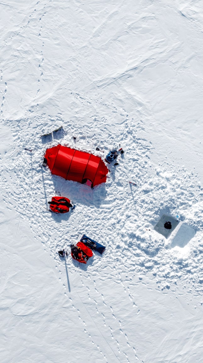 Vue aérienne d'un camp installé sur une vaste étendue de neige. Au centre, une tente rouge se détache nettement sur le blanc immaculé du paysage. Autour, on distingue du matériel d'expédition, notamment deux pulkas rouges, un trou creusé dans la neige, et plusieurs traces de pas. Matthieu, vêtu de bleu, est visible à proximité de la tente, tandis que Heïdi, en tenue orange, s'affaire non loin. La scène illustre un campement polaire temporaire, isolé et entouré d'un décor glacial spectaculaire.