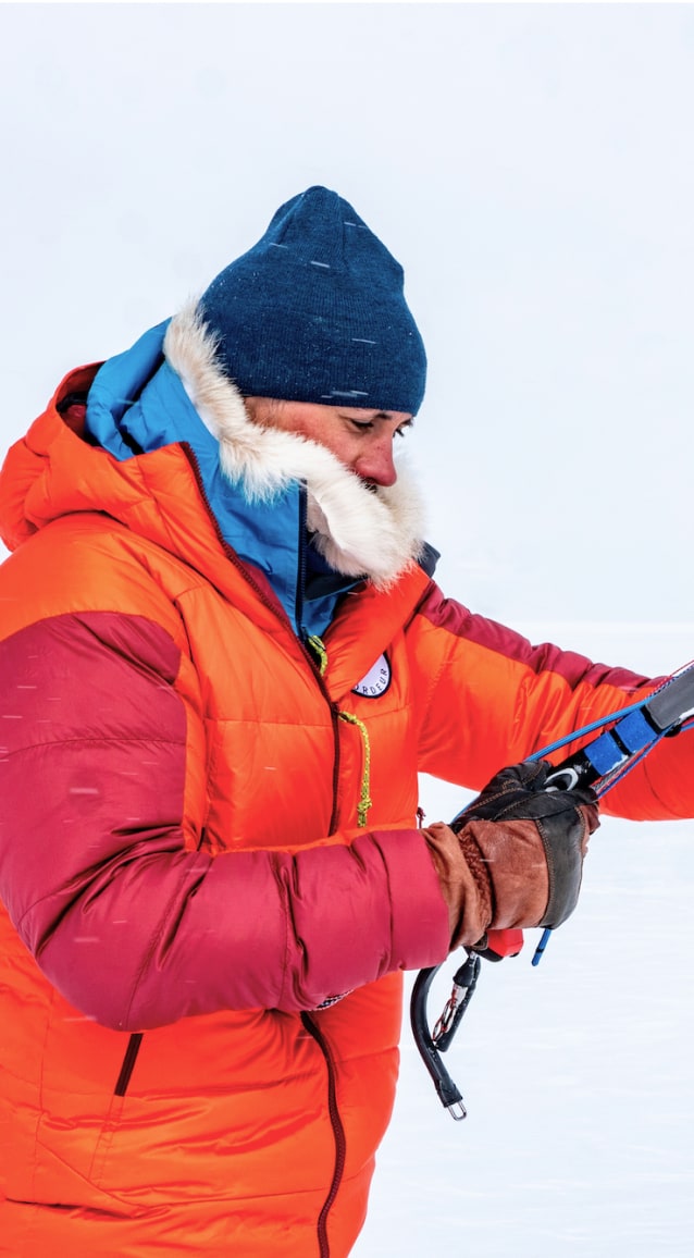 Matthieu, vêtu d'une doudoune orange et rouge, un bonnet bleu et des gants en cuir, ajuste les lignes de contrôle de son aile de traction dans un environnement glacé. Le fond est entièrement blanc, suggérant une surface gelée ou enneigée. Il se concentre sur son geste, illustrant la préparation minutieuse d'une activité de snowkite en conditions polaires.