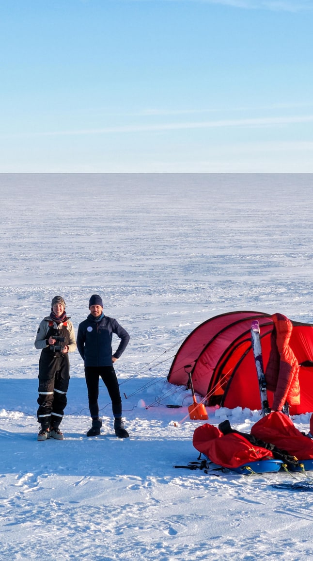 Sur une vaste plaine glacée baignée de lumière, Matthieu et Heïdi se tiennent côte à côte devant une grande tente d'expédition rouge. Ils portent une tenue plus légère, adaptée à un moment de détente après l'effort : Matthieu est en pull et bonnet, tandis qu'Heïdi tient les commandes d'un drone entre ses mains, concentrée sur le pilotage. À leurs pieds, quatre pulkas rouges — servant à transporter les radars utilisés pour les relevés de la calotte polaire — sont alignées près de la tente. Deux paires de skis sont plantées dans la neige, l'une d'elles servant de support à un manteau orange posé en équilibre. À proximité, un panneau solaire capte les rayons du soleil pour recharger le matériel. Une longue corde verte relie les instruments scientifiques étendus au loin sur la glace. Le ciel bleu limpide et la lumière rasante soulignent la quiétude de cette scène d'expédition arctique.