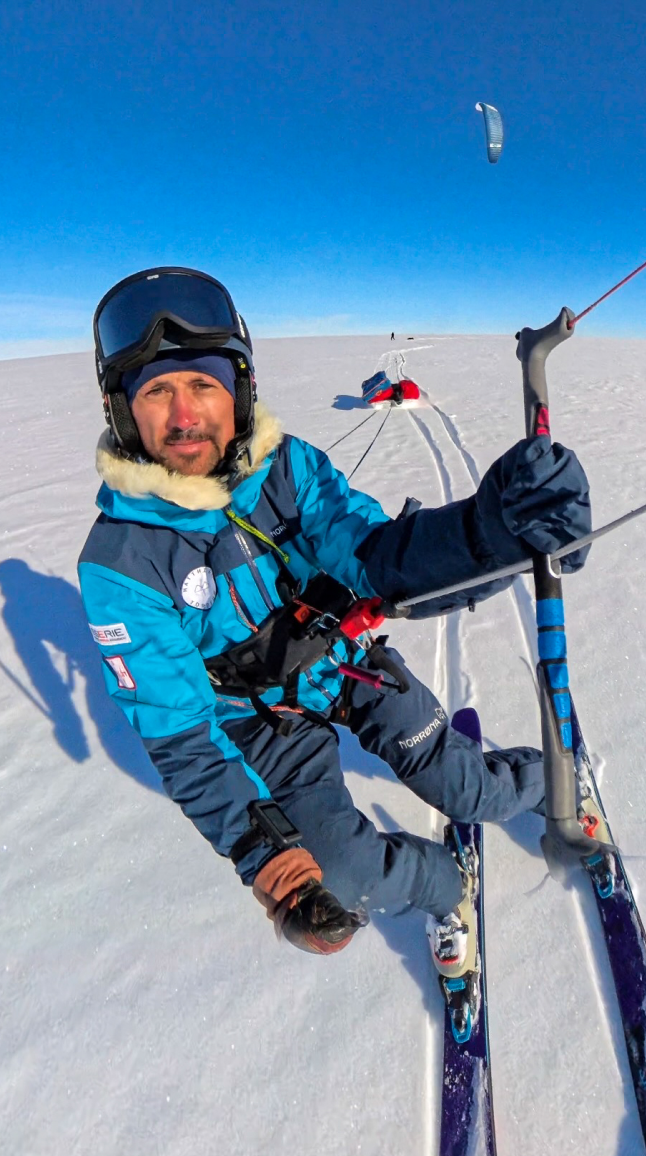 Matthieu pratique le snowkite sur une vaste plaine enneigée. Penché légèrement vers la gauche, les genoux fléchis sur ses skis, il tient fermement la barre de son aile de traction. Sa pulka rouge est tractée derrière lui sur la neige. Il porte une combinaison bleue, un bonnet bleu et un casque sur lequel repose son masque de ski. Son visage exprime un léger sourire. Le ciel est d'un bleu éclatant, sans nuage, et la lumière intense accentue la blancheur du paysage. Au loin, on distingue Heïdi, plus petite dans la perspective, avançant elle aussi sur la neige. L'image transmet une sensation de mouvement, de vitesse et de liberté au cœur d'un environnement polaire infini.