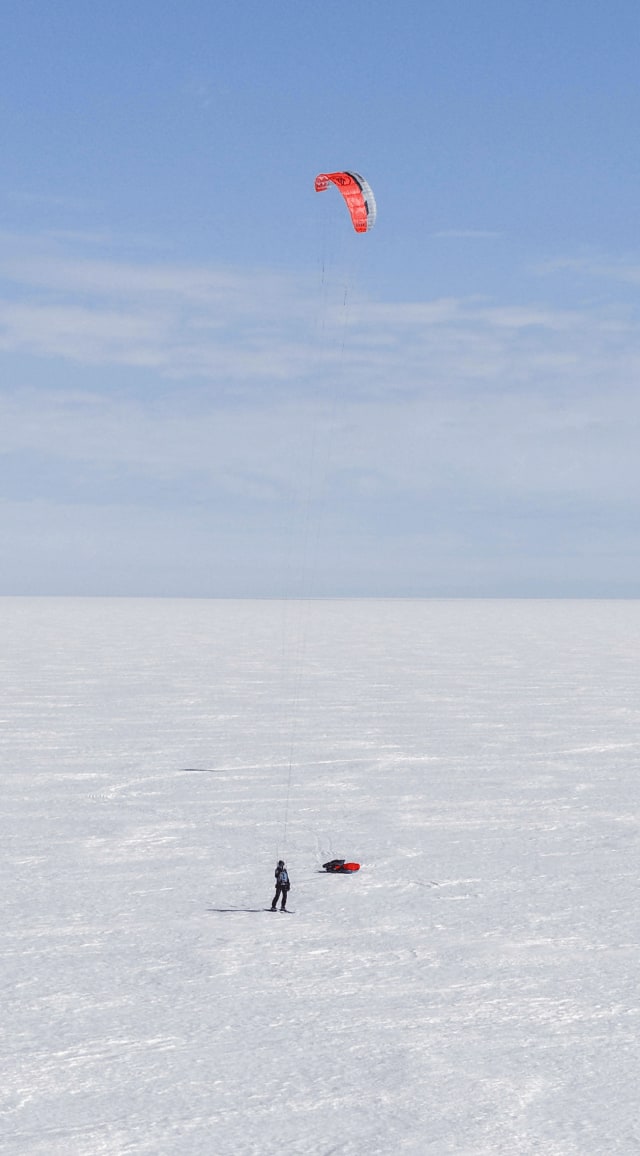 Sur une vaste étendue blanche et immaculée de la calotte polaire, deux explorateurs, Matthieu et Heïdi, avancent en ski tractés par de grandes voiles rouges de kite. Leurs pulkas glissent derrière eux, laissant des traces sinueuses sur la neige. À l'horizon, on aperçoit une structure scientifique isolée, un dôme blanc entouré de quelques équipements, contrastant avec l'immensité du désert de glace. Le ciel, d'un bleu pâle uniforme, se fond à l'horizon dans une lumière douce et silencieuse. 