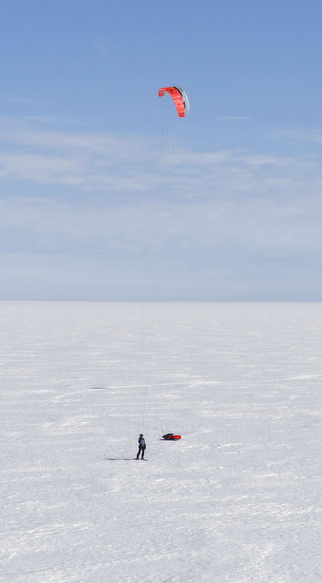 Sur une vaste étendue blanche et immaculée de la calotte polaire, deux explorateurs, Matthieu et Heïdi, avancent en ski tractés par de grandes voiles rouges de kite. Leurs pulkas glissent derrière eux, laissant des traces sinueuses sur la neige. À l'horizon, on aperçoit une structure scientifique isolée, un dôme blanc entouré de quelques équipements, contrastant avec l'immensité du désert de glace. Le ciel, d'un bleu pâle uniforme, se fond à l'horizon dans une lumière douce et silencieuse.