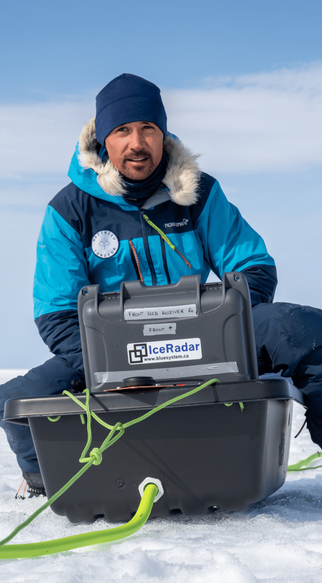 Matthieu est assis sur la neige, au cœur d'un vaste paysage glacé qui s'étend à perte de vue sous un ciel bleu clair légèrement voilé de nuages. Il porte une veste d'expédition bleue et turquoise à capuche bordée de fourrure, un bonnet bleu marine et un pantalon technique isolant. Devant lui, une grande caisse noire portant l'étiquette IceRadar – bluesystem.ca contient le matériel scientifique utilisé pour leurs relevés de la calotte polaire. Une corde verte relie la caisse à d'autres instruments étendus sur la glace, visibles en arrière-plan. Matthieu regarde vers la droite de l'image, le visage concentré et légèrement éclairé par la lumière du soleil arctique. L'ensemble évoque la rigueur et la précision du travail scientifique mené dans des conditions extrêmes.