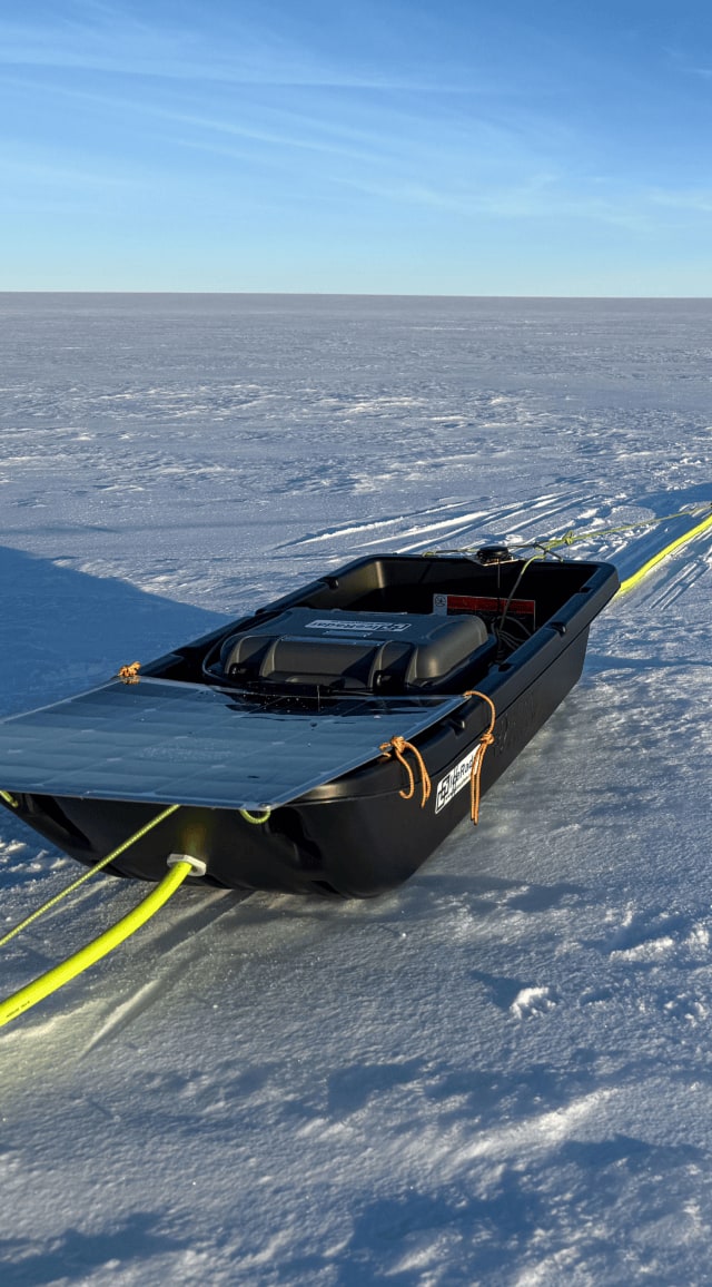 Sur la neige étincelante d'un désert glacé, une longue file d'équipements scientifiques s'étire à perte de vue. Au premier plan, une pulka noire contient un boîtier étiqueté IceRadar – bluesystem.ca ainsi qu'un panneau solaire fixé à l'arrière, incliné vers le soleil pour capter l'énergie. Une corde fluorescente jaune relie cette pulka à d'autres modules similaires alignés en série sur la calotte polaire. Le ciel dégagé, d'un bleu clair, se fond à l'horizon sans aucune aspérité. L'image dégage une impression de silence absolu et d'immensité, témoignant du travail scientifique mené dans un environnement isolé et extrême.