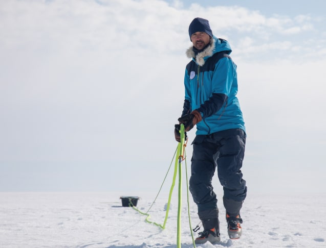 Matthieu avance sur la calotte glaciaire, tirant derrière lui une corde fluo reliée à une pulka noire contenant le matériel scientifique. Il porte une combinaison bleue et un bonnet sombre, avec des gants en cuir et des chaussures de ski. Le vent semble léger, le ciel clair et la lumière rasante souligne le relief du sol enneigé. La scène traduit à la fois l'effort physique et la concentration nécessaire pour la progression dans cet environnement polaire.