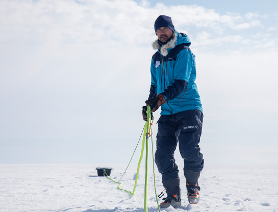 Matthieu avance sur la calotte glaciaire, tirant derrière lui une corde fluo reliée à une pulka noire contenant le matériel scientifique. Il porte une combinaison bleue et un bonnet sombre, avec des gants en cuir et des chaussures de ski. Le vent semble léger, le ciel clair et la lumière rasante souligne le relief du sol enneigé. La scène traduit à la fois l'effort physique et la concentration nécessaire pour la progression dans cet environnement polaire.