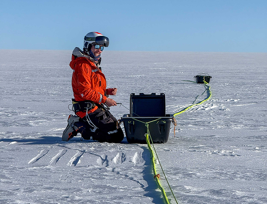 Sur la calotte glaciaire, Heïdi est agenouillée dans la neige, souriante face à la caméra. Elle porte une veste orange vif, un casque et des lunettes de ski, avec un baudrier d'expédition autour de la taille. Devant elle, une mallette scientifique est ouverte, reliée à un long câble jaune fluorescent qui serpente sur la glace jusqu'à une autre caisse plus loin. La lumière met en valeur la blancheur du paysage et l'énergie positive du moment.